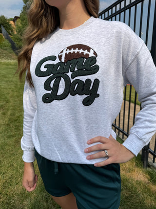 A woman wearing a light heather gray long-sleeve sweatshirt with a 'Game Day' football graphic, paired with dark green athletic shorts, standing outdoors on a grassy field near a black fence.