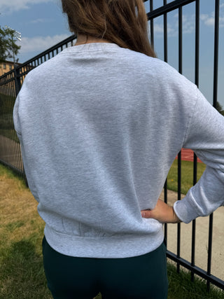 A person viewed from behind wearing a light heather gray long-sleeve crewneck sweatshirt and dark teal leggings, standing outdoors next to a black metal fence.