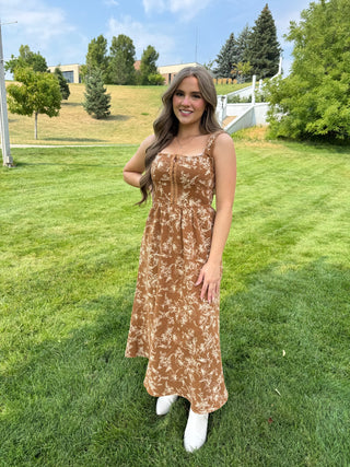 A woman wearing a brown and beige floral print maxi dress with white boots, standing on a green grass lawn outdoors.