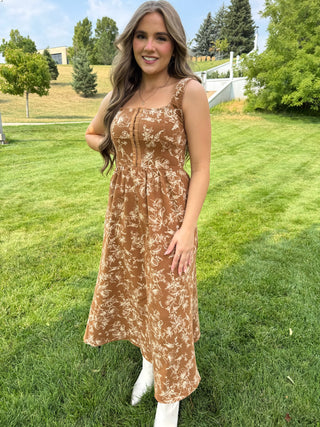 A woman wearing a brown and beige floral print midi dress with white boots, standing on a green grass lawn outdoors.