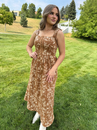 A woman wearing a brown and beige floral print midi dress stands on a green grassy lawn outdoors.