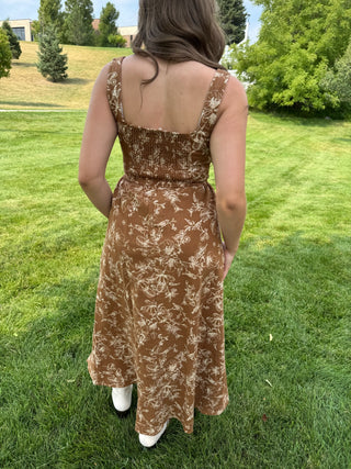 A woman viewed from behind wearing a brown sleeveless maxi dress with a beige floral pattern, standing on a green grass lawn.