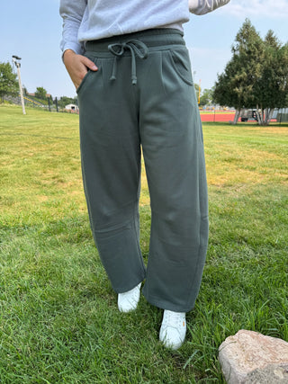 A person standing outdoors on grass wearing loose, wide-leg drawstring sweatpants in a muted sage green color, paired with a light gray sweatshirt and white sneakers.