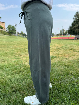 A person standing on a grassy field wearing loose, wide-leg sweatpants in a muted sage green color, paired with white sneakers. The photo is taken from behind, focusing on the fit and drape of the pants.