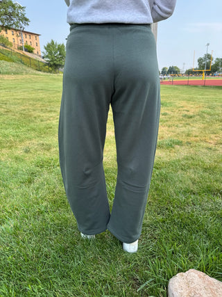 A person standing outdoors on a grassy field wearing loose, wide-leg sage green sweatpants, a light gray sweatshirt, and white sneakers.