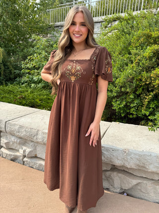A woman wearing a brown midi dress with floral embroidery on the bodice and sleeves, standing outdoors in front of a stone wall and green bushes.