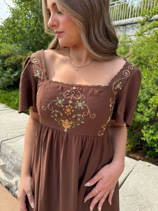 A woman wearing a brown short-sleeve dress with floral embroidery on the bodice, standing outdoors on a stone walkway with green foliage in the background.