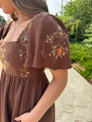 A woman wearing a brown dress with floral embroidery on the bodice and sleeves, photographed outdoors on a stone path with green foliage in the background.