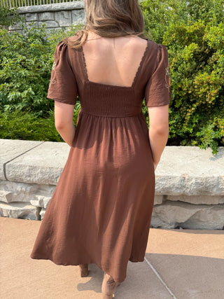 A woman wearing a brown midi dress with puff sleeves and a smocked back, photographed from behind in an outdoor garden setting.