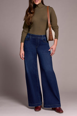 Person wearing a green turtleneck and blue jeans with a brown bag on a neutral background